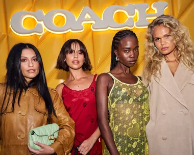 Four women posing in front of a yellow background with the illuminated Coach logo, wearing fashionable outfits.